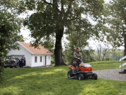 Sieviete uz traktora dārzā pļaut zālienu, fonā māja